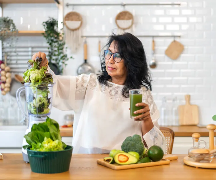 A woman in a kitchen blending leafy greens while holding a green juice, representing the benefits of supplements for cleanse and detox.